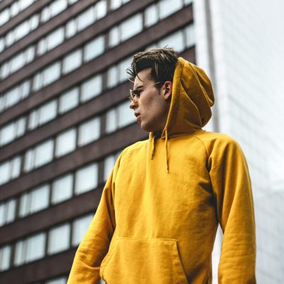 Stylish man poses in yellow hoodie against urban building backdrop.